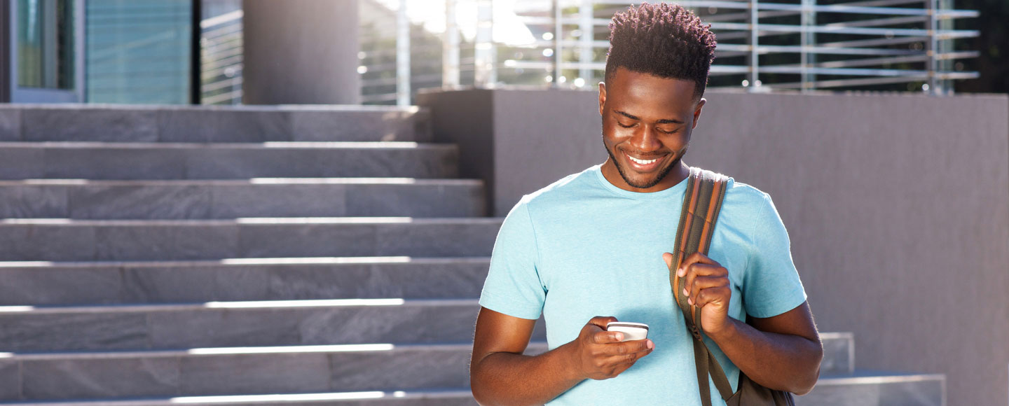 Photo of a young college student, standing on the steps of a library or other campus building, holding his phone and smiling.