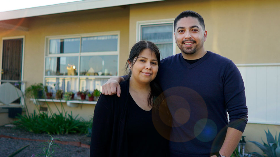 A woman and man stand next to each other and smile at the camera. The man has his arm around the woman. They stand in front of a house.
