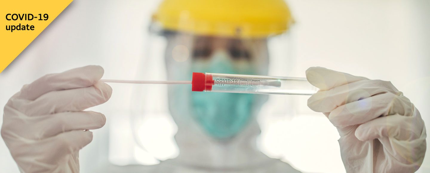 Person in white lab coat and protective headgear (blurred in background) looks closely at a COVID-19 testing swab going into a container.