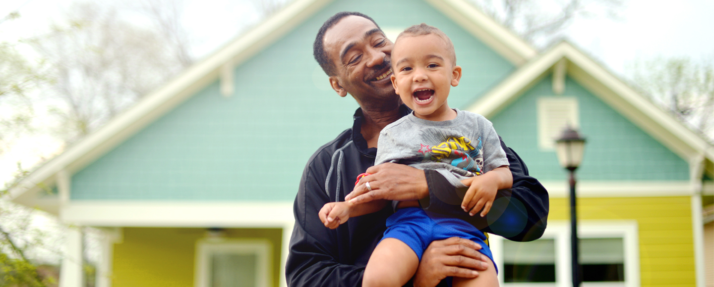 A smiling man holds a laughing boy in front of a house.