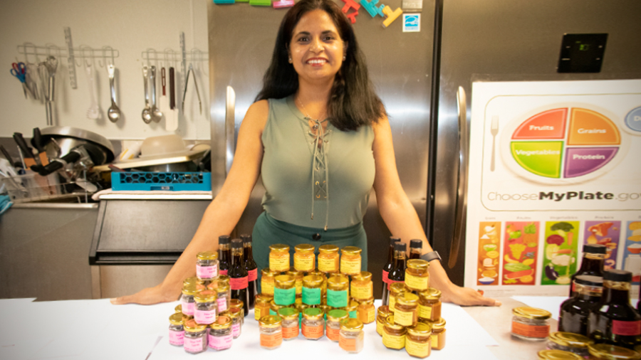 Ranjana Hans smiles as she stands behind a table in a kitchen. On the table are stacked spices. Behind her is a refrigerator and sink with kitchen utensils above it.
