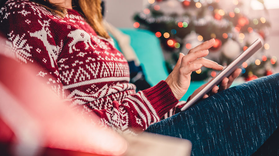 A person wearing a holiday sweater sits in their living room at home and uses their tablet. In the background is a Christmas tree.