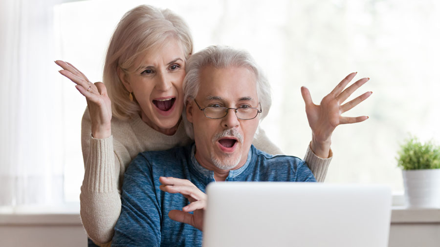 A man sits in front of the computer and his wife stands behind him. They both look at the screen and show expressions of delight at a deal they just found online.