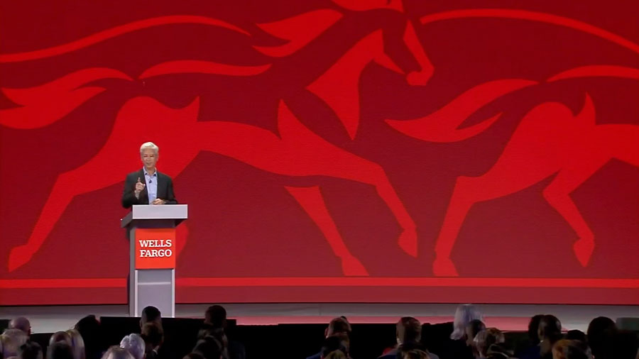 Wells Fargo CEO Charlie Scharf stands behind a lecturn to address an audience of employees. A red silhouette of horses is in the background behind him on stage.