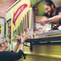 A bearded man in a black T-shirt serves food to female customers through the window of a yellow food truck.