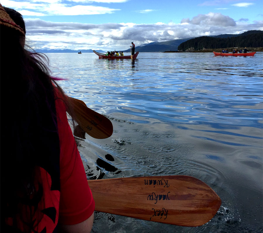 People are sitting in a yaakw and holding paddles with their backs turned to the camera. On the paddle, the words 'Kéex’ yaakw Kwáa' are visible. Other yaakws are on the water in the distance.