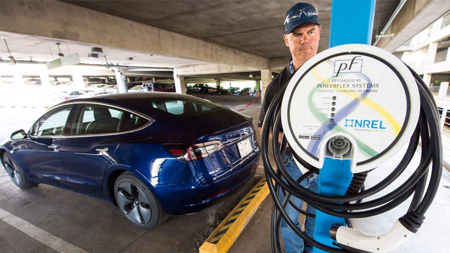 A car is parked next to an electric vehicle charging station at the NREL parking garage. A man stands behind the charging station.